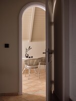 Vista interna di una stanza con soffitto alto, una zona pranzo con sedie in vimini e un tavolo, e una porta che conduce in un'altra stanza.