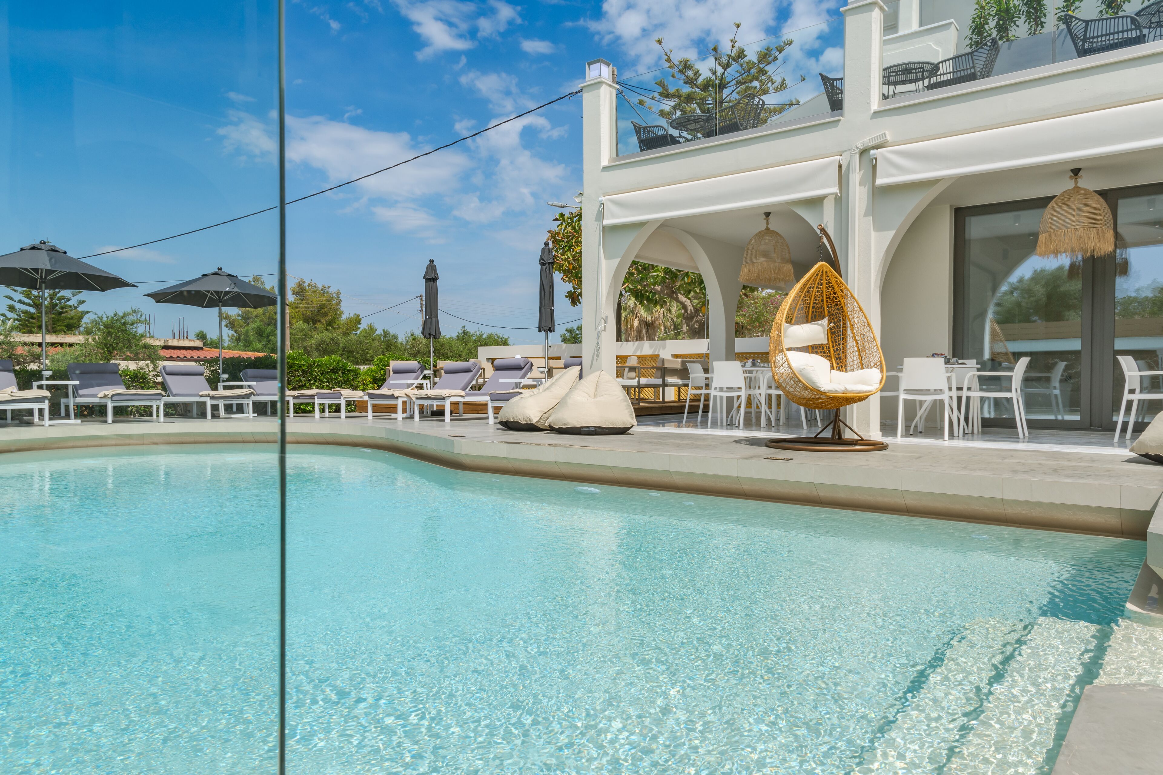 Un área de piscina con sillones de descanso y una hamaca, rodeada por un edificio blanco con amplias ventanas y asientos al aire libre.