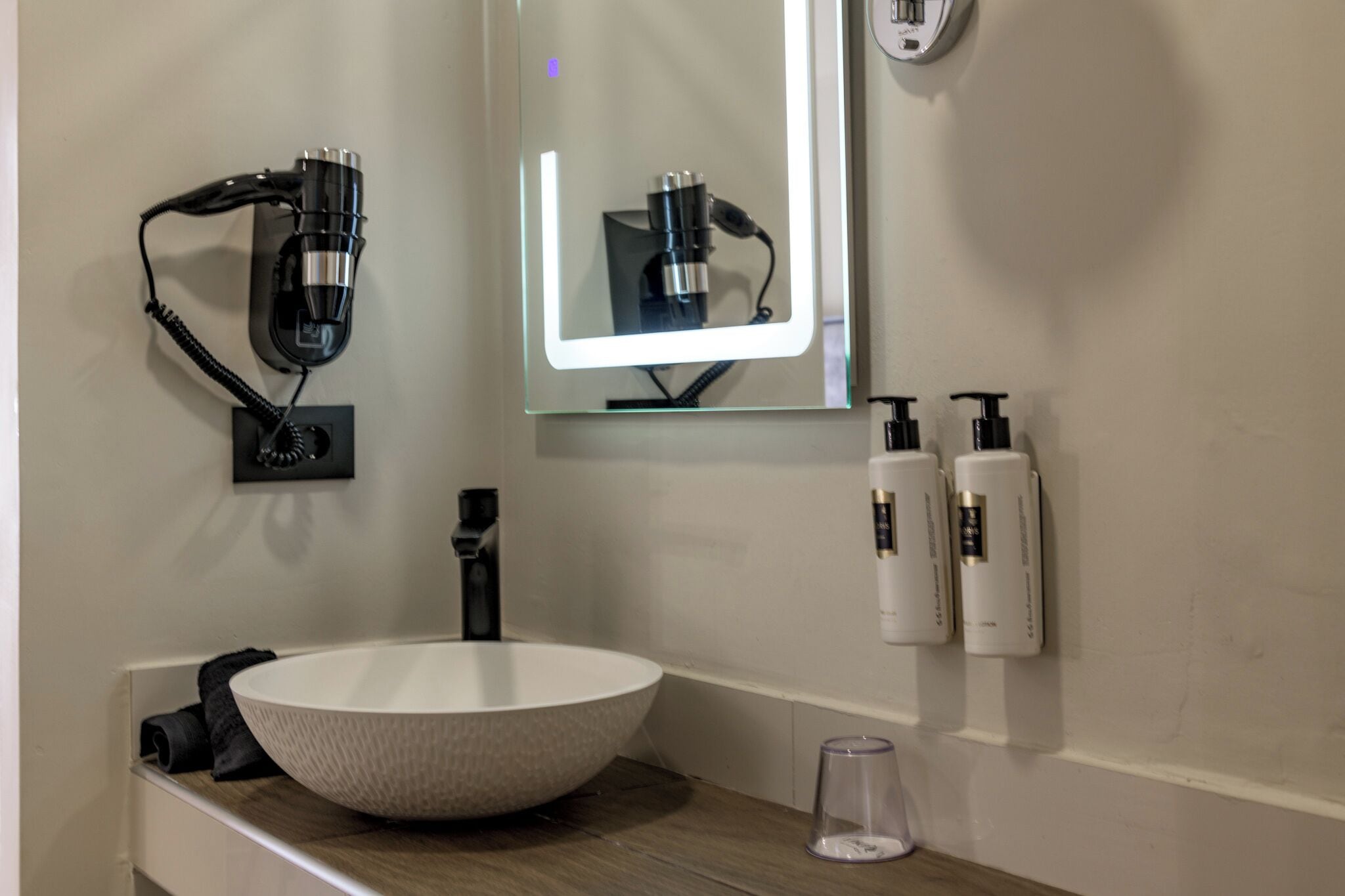 A bathroom with a large white sink, a mirror with a light, and two bottles of shampoo and conditioner on the wall.