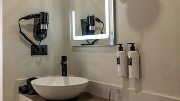 A bathroom with a large white sink, a mirror with a light, and two bottles of shampoo and conditioner on the wall.