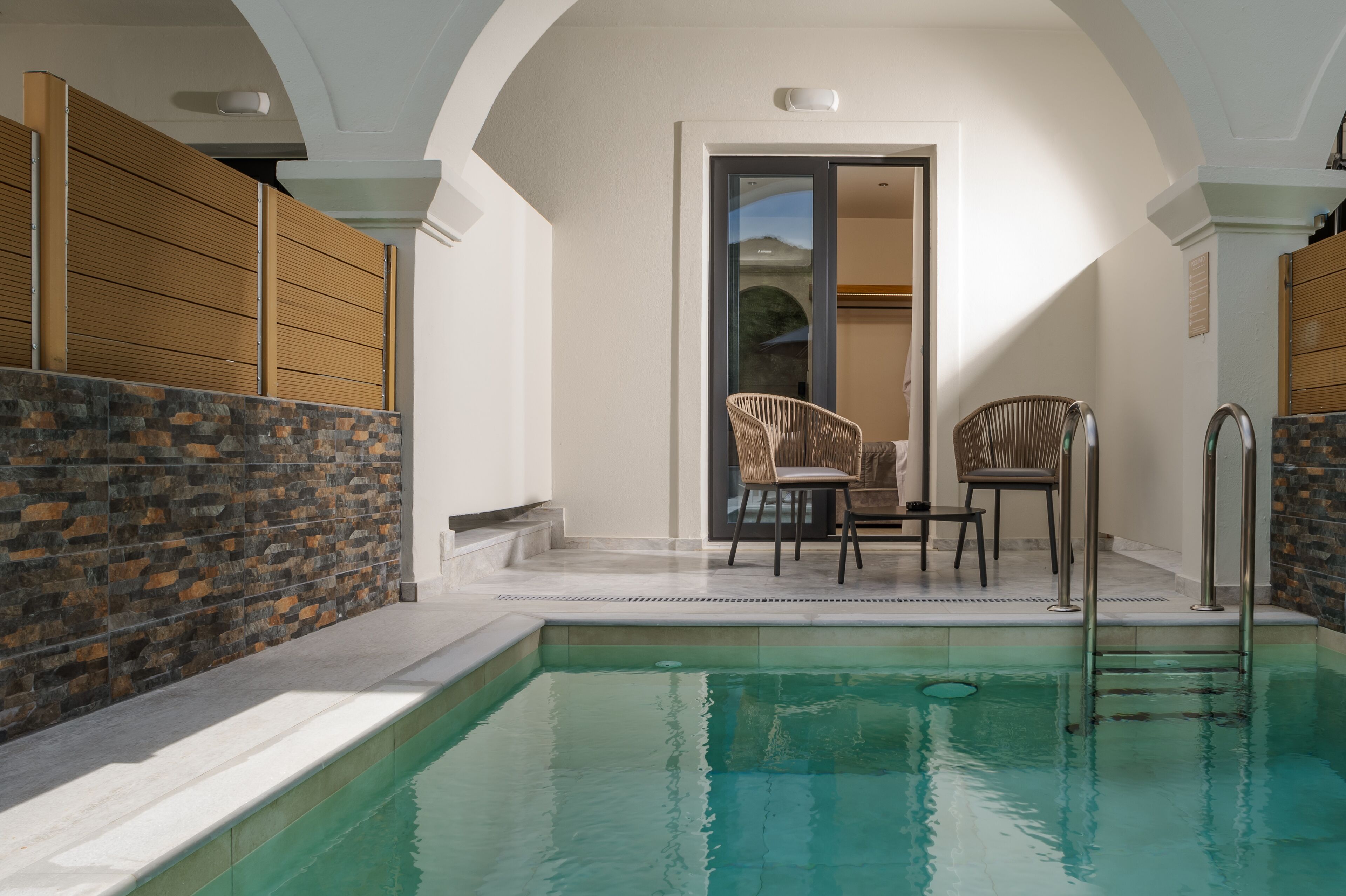 Una piscina con muro de piedra y paneles de madera, dos sillas de mimbre y una puerta que da a una habitación.