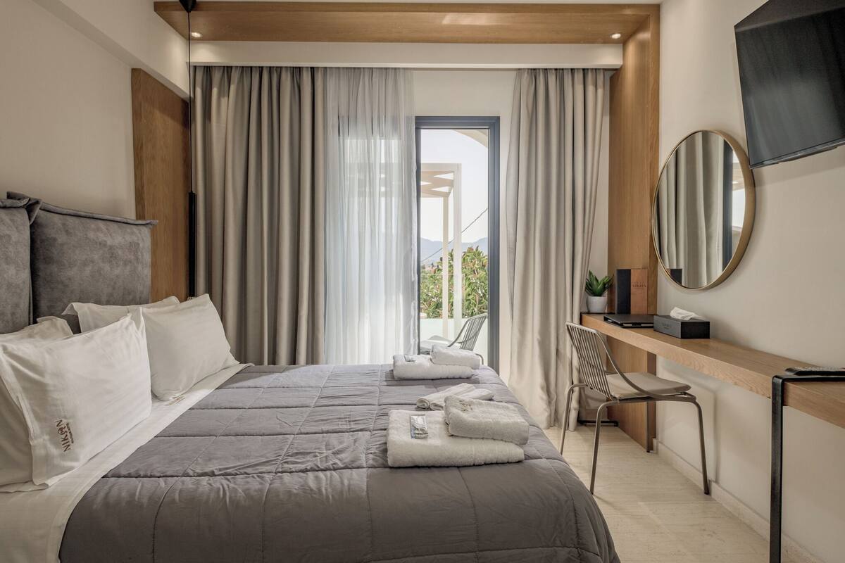 A modern hotel room with a large bed, a desk, and a chair. There is a window with curtains, a TV mounted on the wall, and a potted plant on the desk.