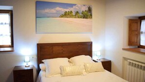 Une chambre à coucher avec une tête de lit en bois, un lit recouvert de linge de lit blanc, deux lampes de chevet et un tableau représentant une scène de plage accroché au mur.