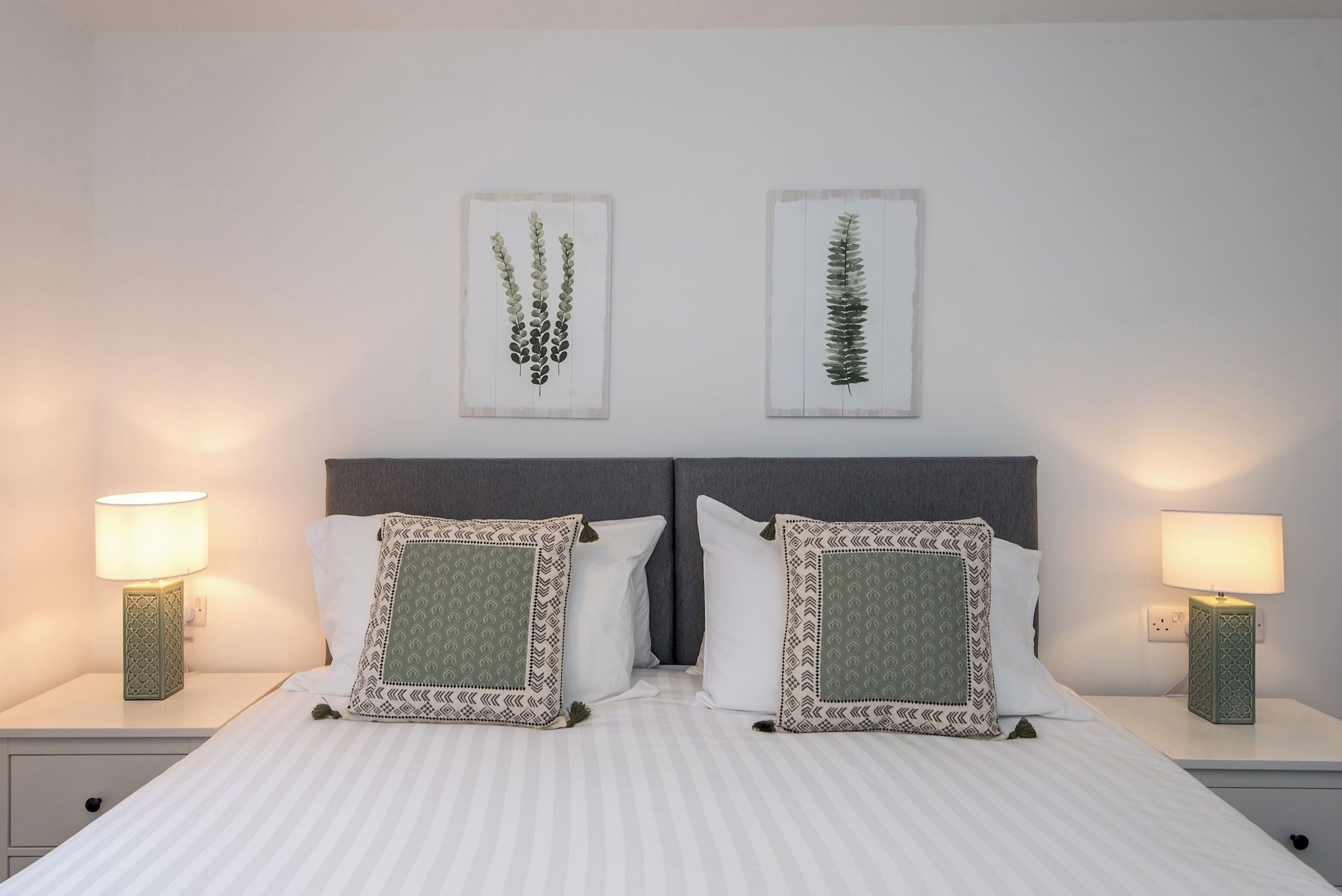A double bed with a grey headboard, two bedside tables with lamps, and two wall-mounted plant prints.