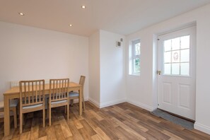 Een eethoek met een houten tafel en stoelen, een witte deur en een raam met uitzicht op groen.