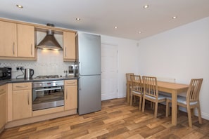 Cocina compacta con armarios de madera, un refrigerador de acero inoxidable, una estufa a gas y una mesa de comedor con sillas.