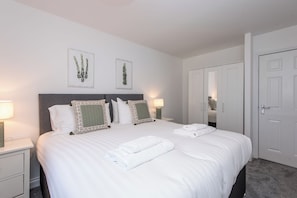 Een netjes opgemaakt bed met witte lakens en twee kussens, een nachtkastje met een lamp, en een deur aan de rechterkant.