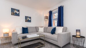 Un salon moderne avec un canapé gris, une table basse en verre et une lampe sur une table d’appoint.