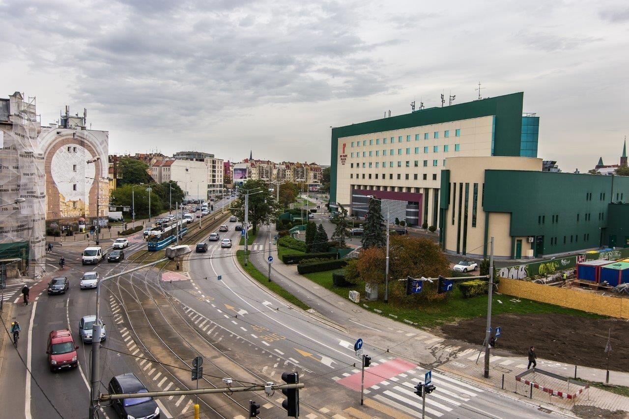 A multi-lane road with vehicles, a pedestrian crossing, and buildings including a large clock tower and a modern building with a green roof.