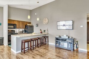Een keuken met houten kasten, een marmeren eiland met barstoelen, een klok en een televisie aan de muur.