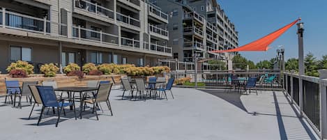 Une terrasse extérieure avec des tables et des chaises, entourée d’une clôture et protégée par une voile d’ombrage rouge.