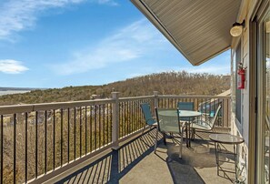 A balcony with a view of a forested area and a body of water. There are two tables and four chairs.