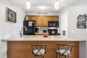 A kitchen with wooden cabinets, a black refrigerator, a microwave, a stove, and two bar stools. - Lake View Condo, Spacious & Only 1 Mile to Silver Dollar City – 1204 (Branson)