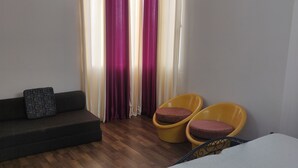 Een kamer met een donkergrijze bank, twee gele stoelen met kussens, een bijzettafeltje en een raam met gordijnen.