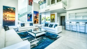A spacious living area with white sofas, a glass coffee table, and a blue rug. There is a staircase leading to an upper level, and a kitchen area with bar stools. - Modern and Fresh Oceanfront Villa (Punta Cana)