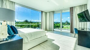 A modern hotel room with a large bed, a desk, and a TV. It has a balcony with a view of greenery and mountains. - Modern and Fresh Oceanfront Villa (Punta Cana)