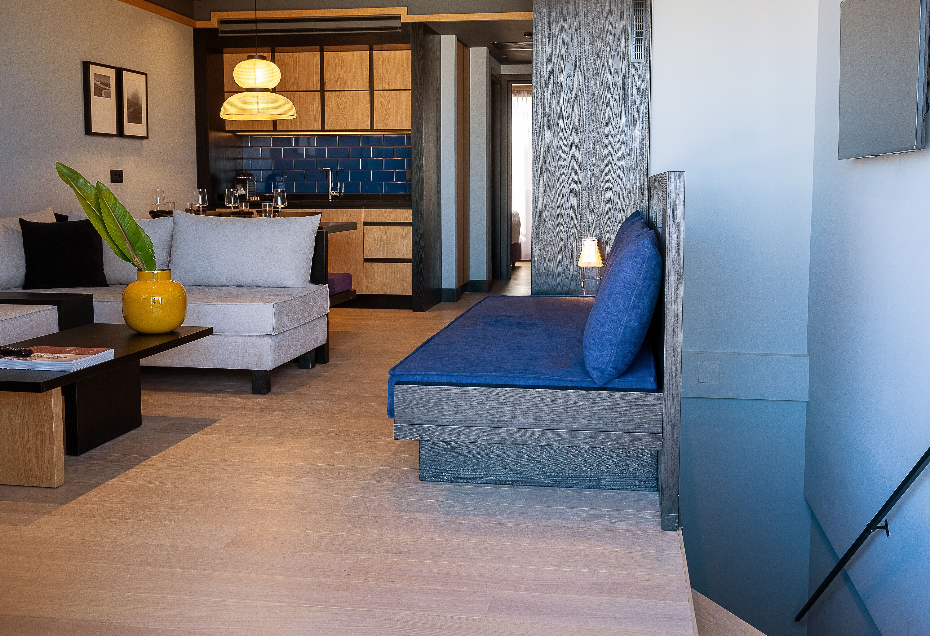 A modern living room with a sofa, a coffee table, and a yellow vase. There is a kitchen area with wooden cabinets and a blue backsplash.
