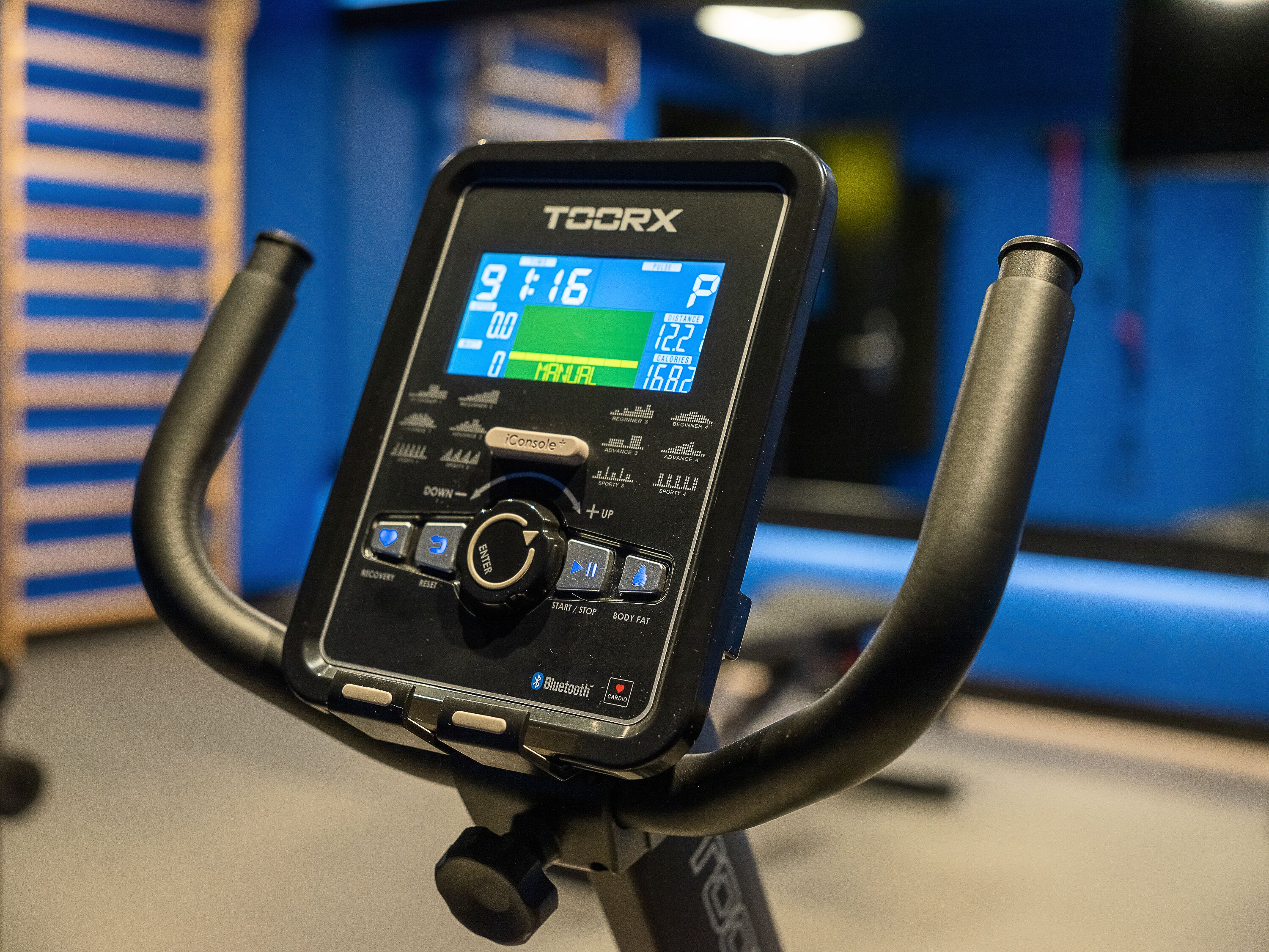 Ein stationäres Fahrrad mit digitaler Anzeige und Bedienfeld, einschließlich Tasten für verschiedene Funktionen und einem Bluetooth-Symbol.