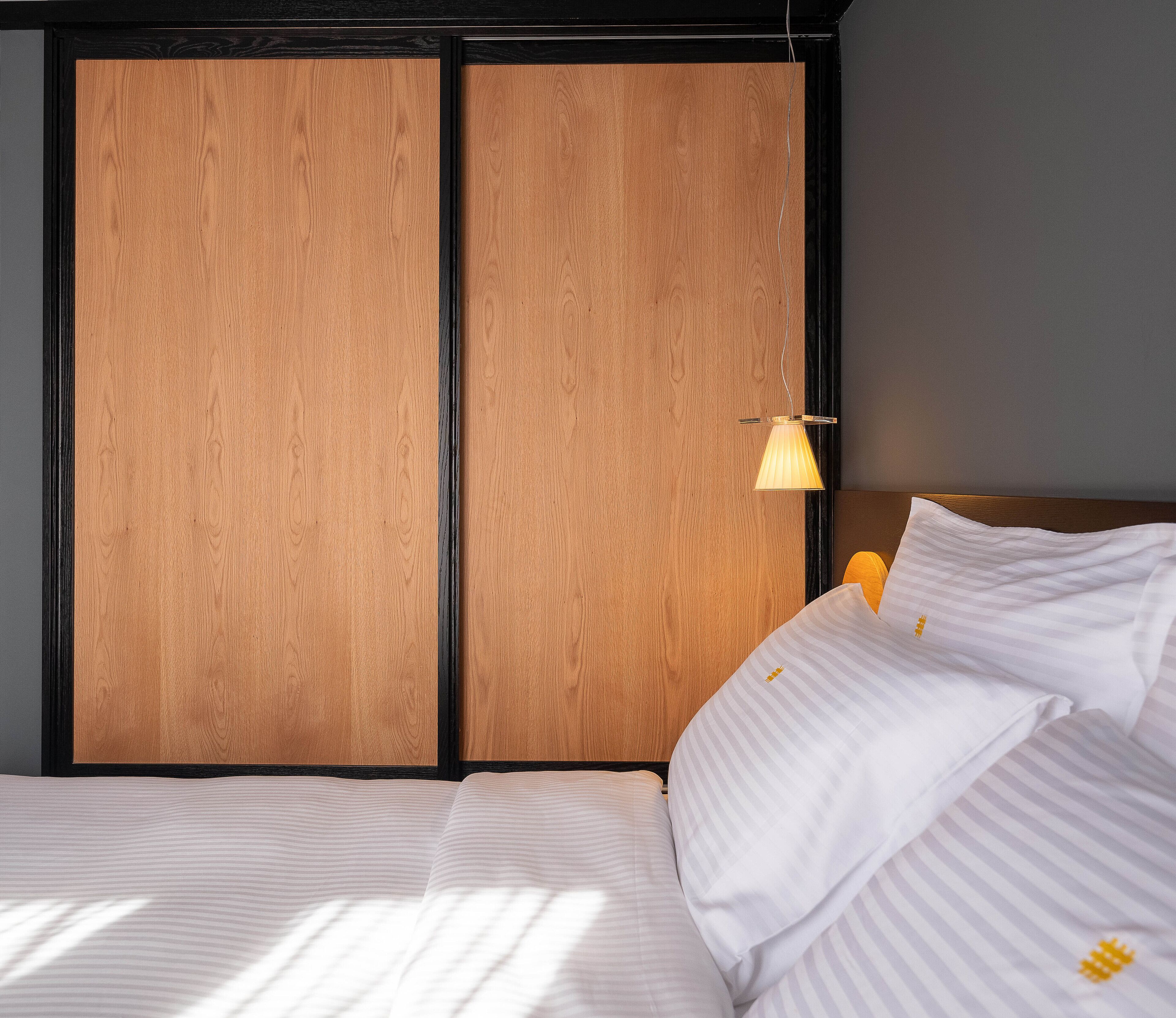 Una cama bien hecha con sábanas blancas y un almohadón a rayas, un cabecero de madera y una lámpara colgante.
