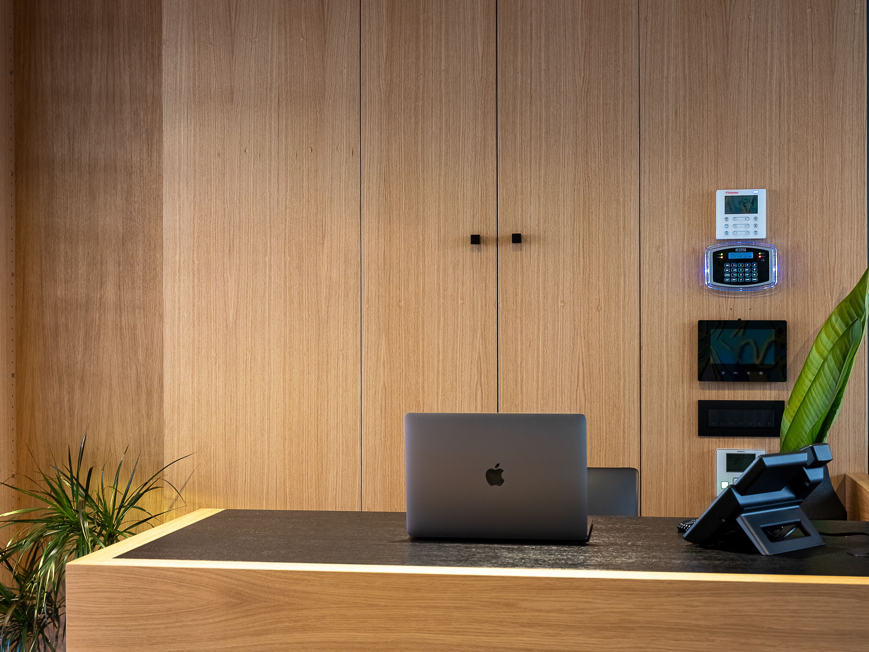 Un bureau en bois avec un ordinateur portable, un clavier numérique pour le système de sécurité, une horloge numérique et une plante.