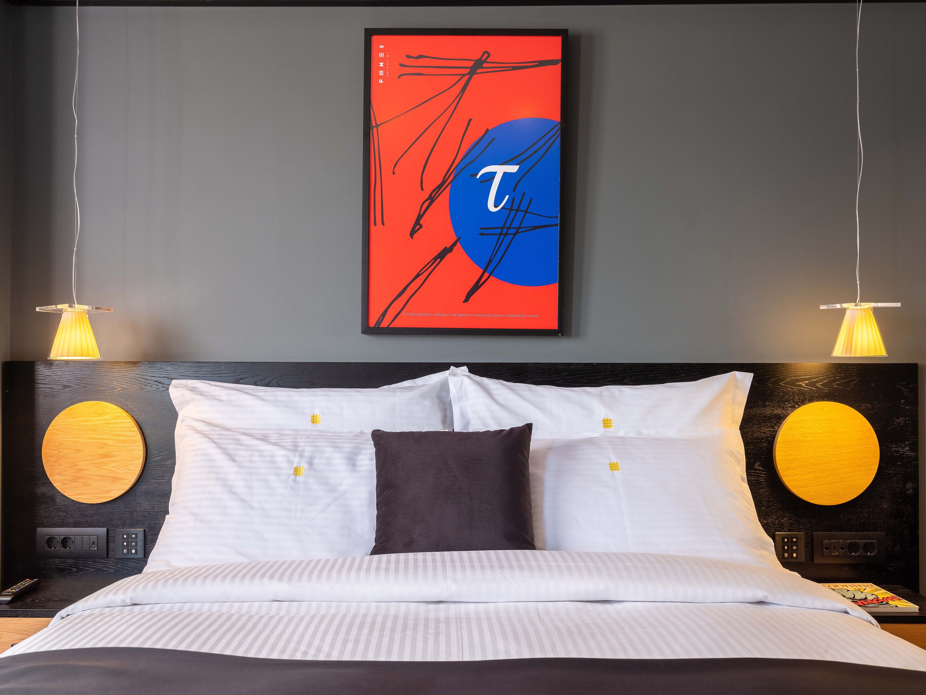 A modern hotel room with a large bed, two bedside tables, a wall-mounted artwork, and two pendant lights.