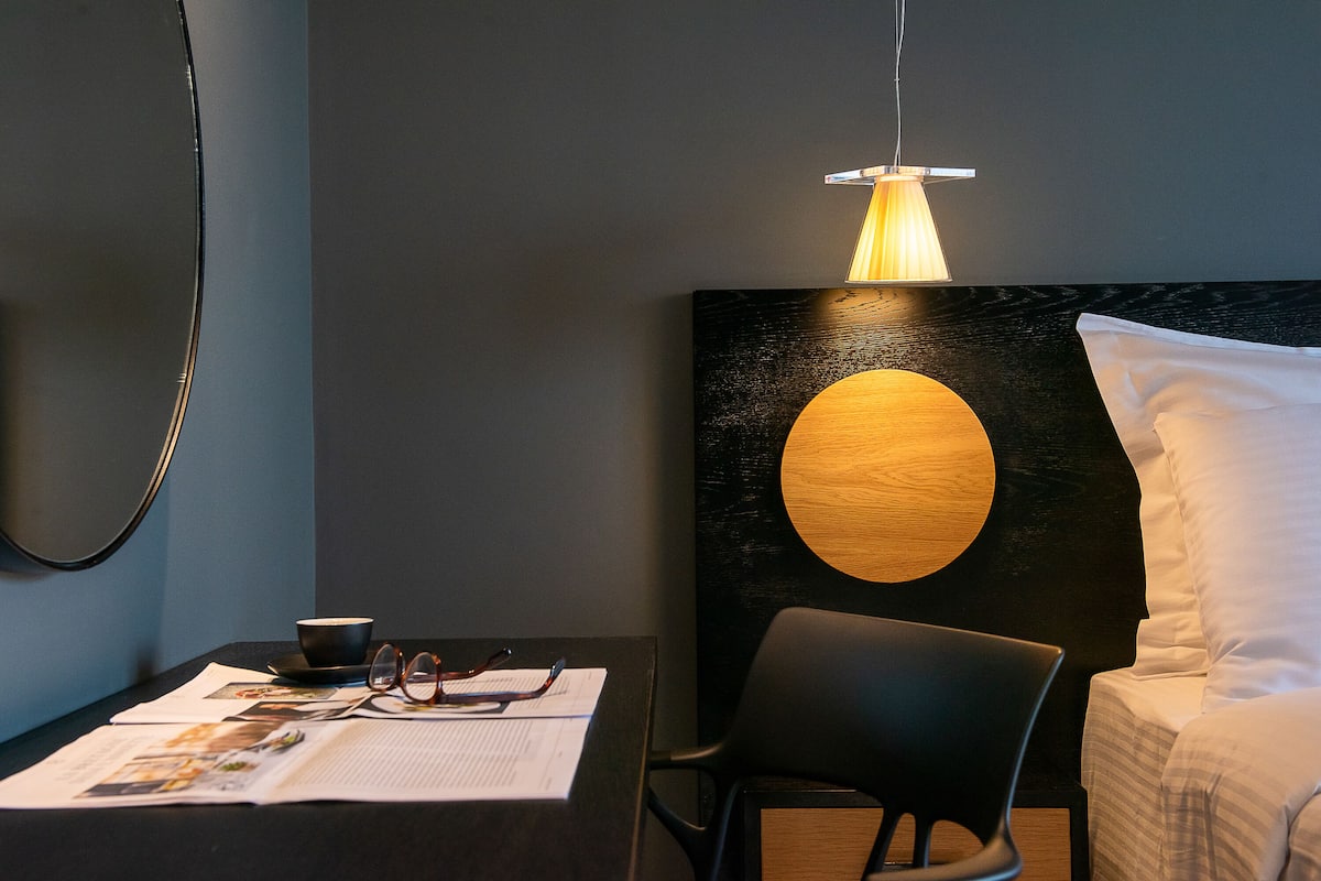 A modern hotel room with a dark wall, a round wooden accent, a black chair, a white bed, and a table with reading materials.