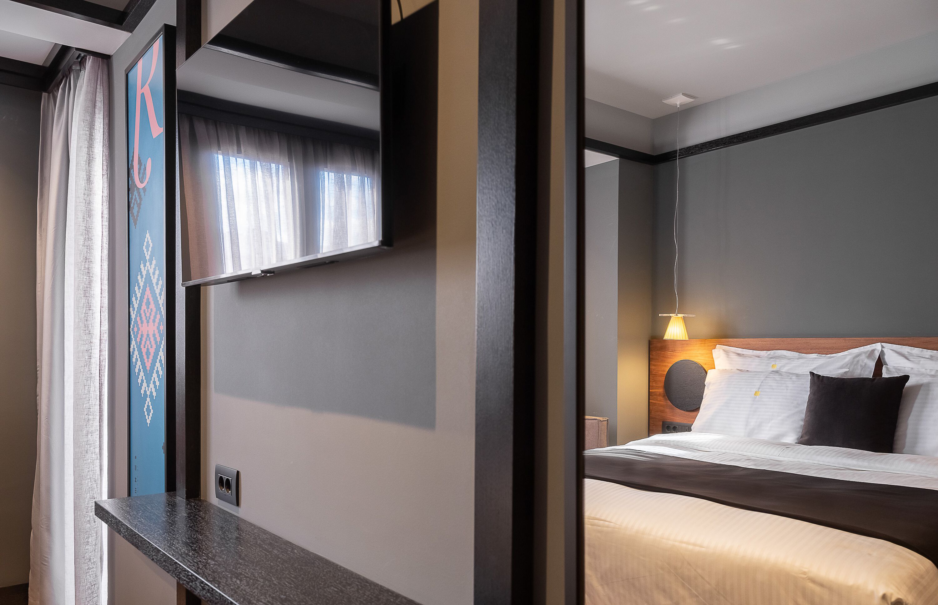 Habitación de hotel con cama, mesita de noche, televisor montado en la pared y ventana con cortinas.