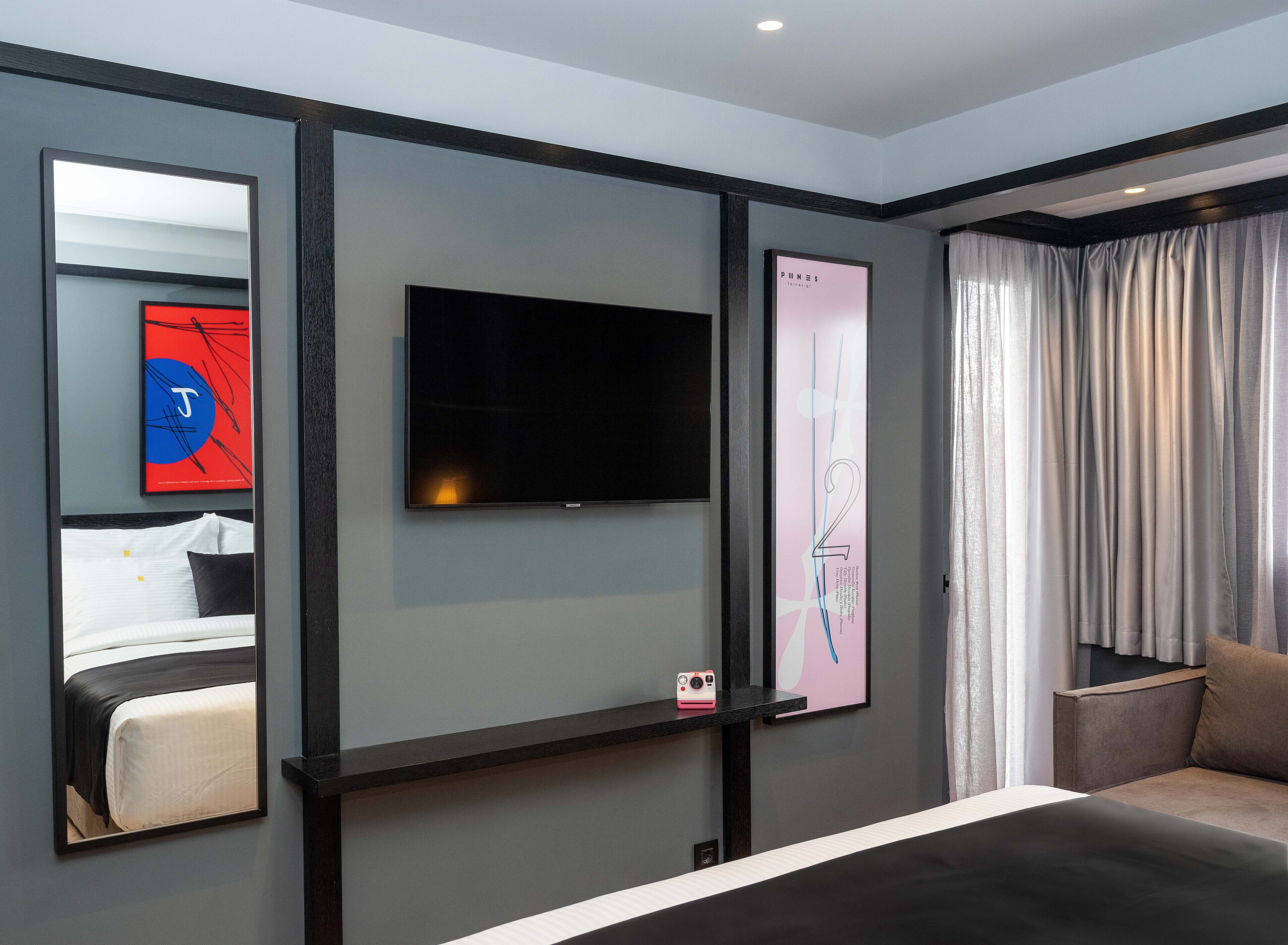 A modern hotel room with a large flat-screen TV mounted on the wall, a bed with white linens, a small bedside table with a pink camera, and a large window with sheer curtains.