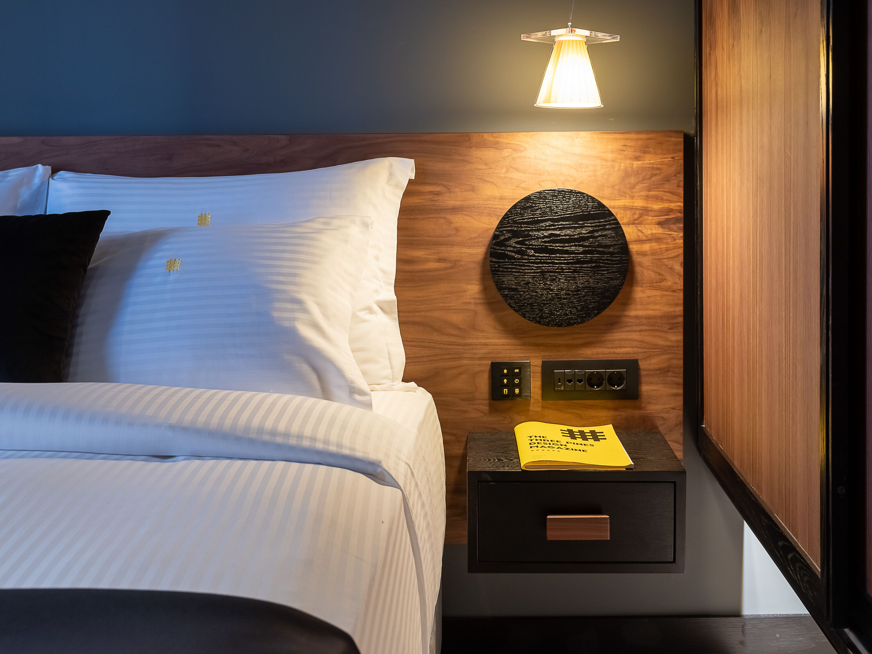 A neatly made bed with white linens and a dark pillow, a round wall decoration, a nightstand with a drawer, and a hanging lamp.