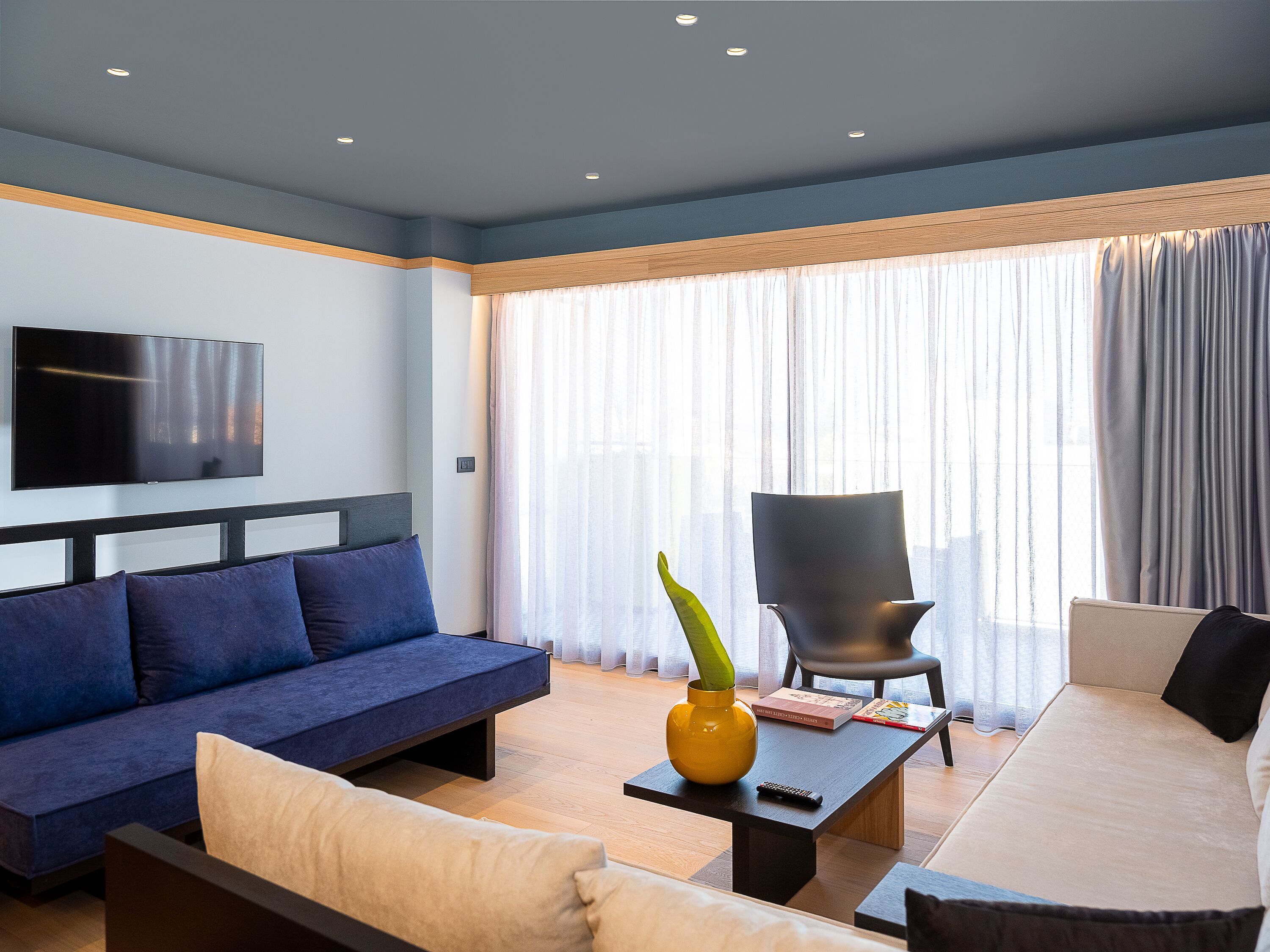A modern living room with a blue sofa, a beige sofa, a black armchair, and a coffee table with a yellow vase and remote control.