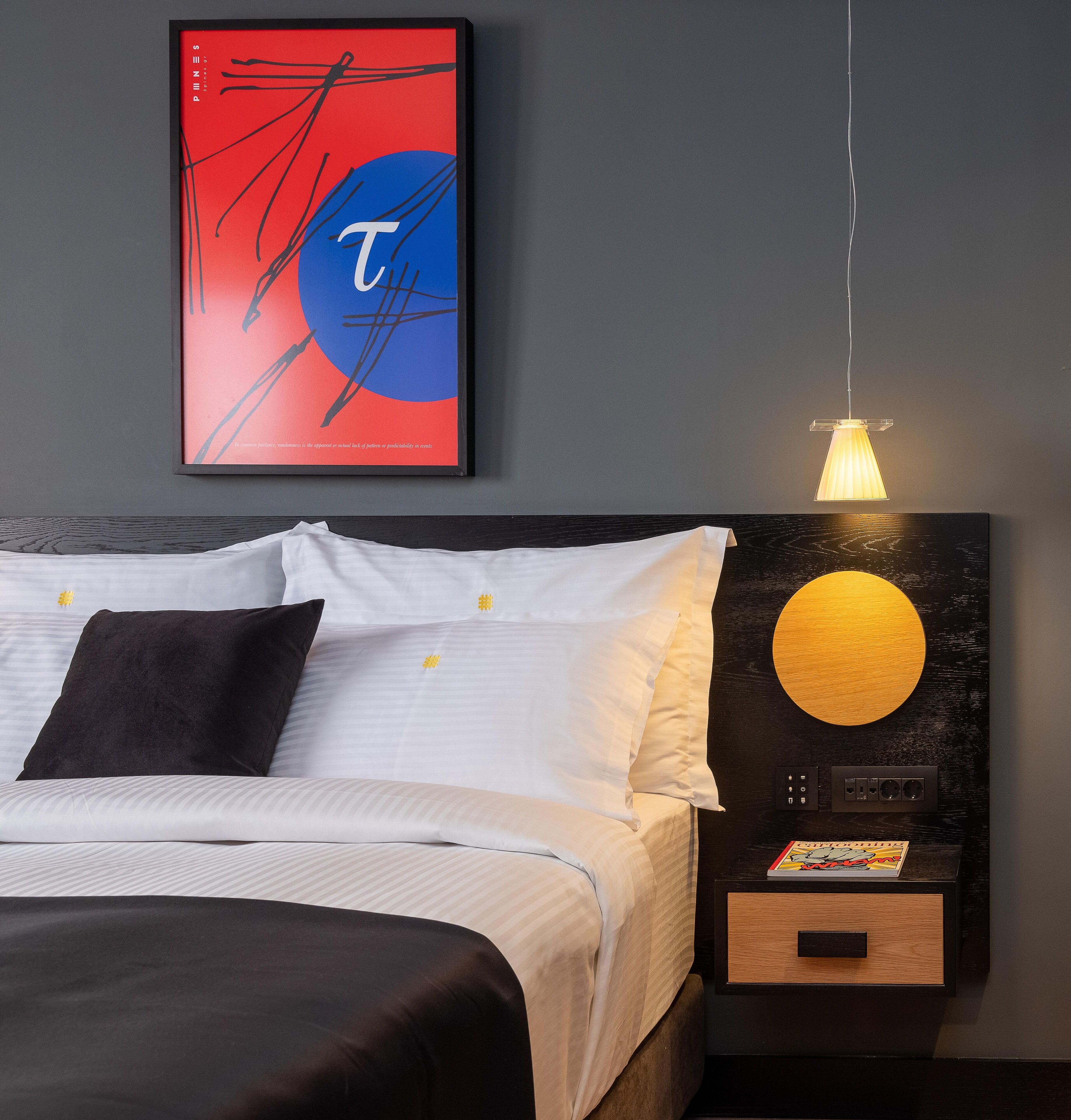 Quarto moderno com cama, roupa de cama branca, travesseiro preto, obra de arte abstrata emoldurada, luminária fixada na parede e mesa de cabeceira com um livro e uma gavetinha.
