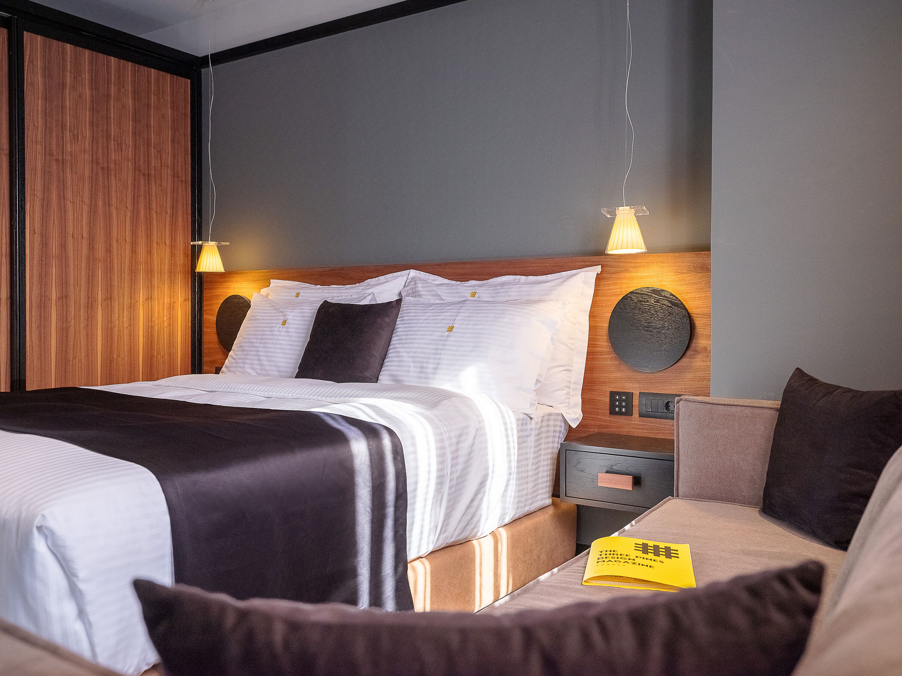 Una cama bien hecha con sábanas blancas y un cojín de color oscuro, un cabecero de madera, una mesita de noche con una lámpara y una revista en el buró.