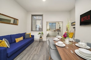 A modern living room with a blue sofa, a dining table set for four, and a kitchen in the background. - Stone Pit Apartments (Warrington)