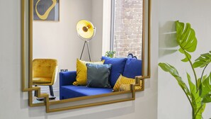 Een kamer met een blauwe bank, gele en zwarte kussens, een gele stoel, een vloerlamp, een inlijstbaar schilderij aan de muur en een plant.