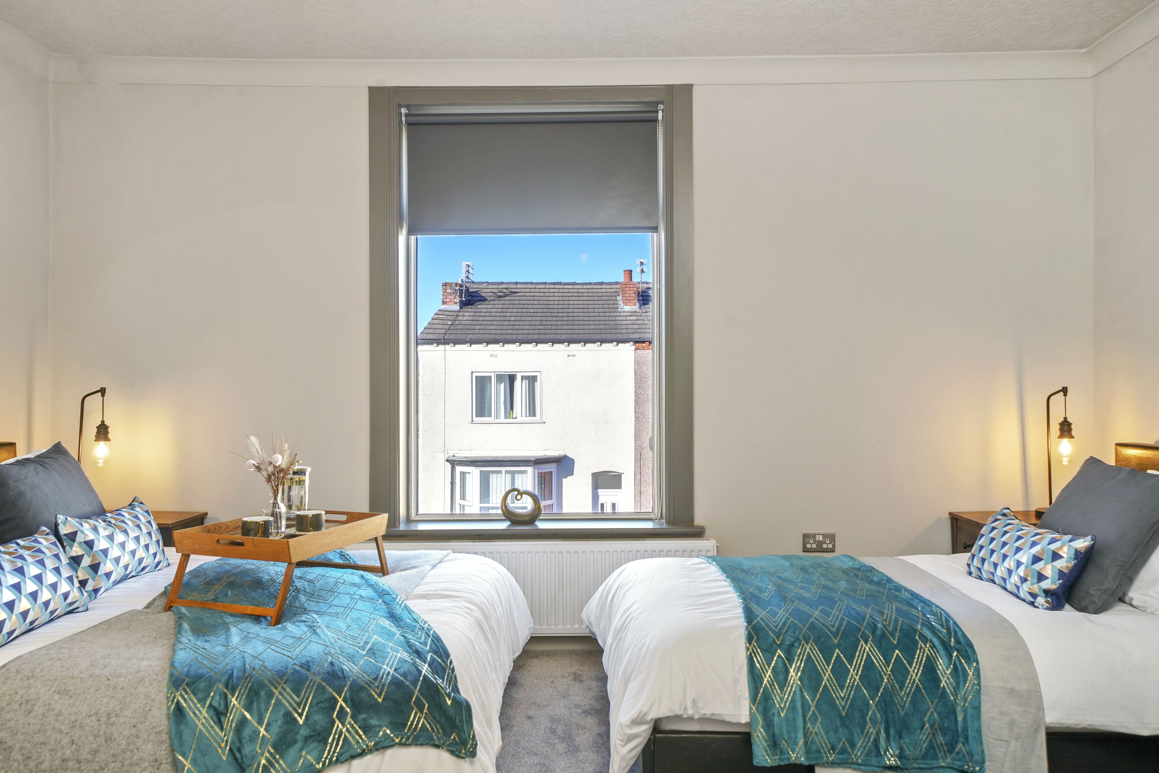 A bedroom with two beds, a window with a view of a building, bedside tables with lamps, and a tray with decorative items.