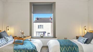 A bedroom with two beds, a window with a view of a building, bedside tables with lamps, and a tray with decorative items.