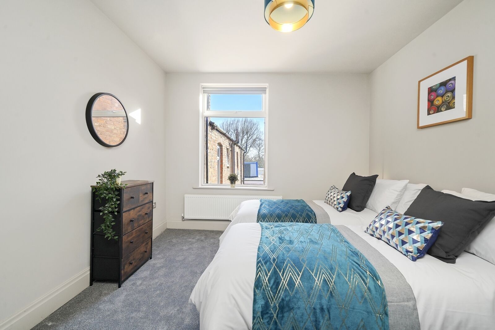 Une chambre avec deux lits, une commode, un miroir, un radiateur et une fenêtre donnant sur des immeubles.
