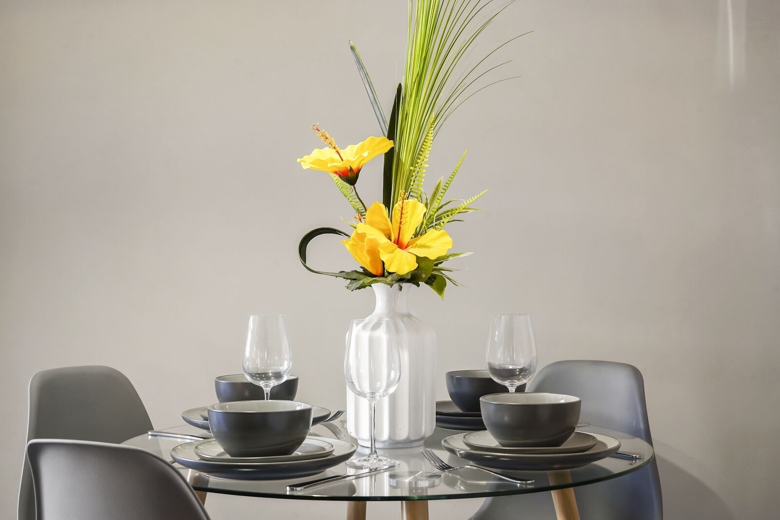 A dining table with a clear glass vase containing yellow flowers and green foliage, surrounded by black bowls and wine glasses.