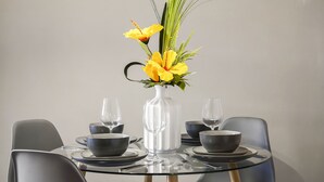 Een ronde glazen tafel met een witte vaas gevuld met gele bloemen en groene bladeren, omringd door zwarte kommen en glazen wijnglazen.