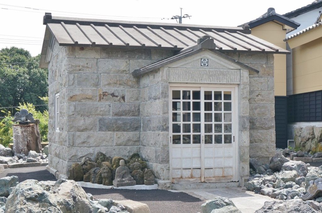 Un edificio de piedra con techo de tejas, una puerta blanca y un pequeño jardín con rocas y una estatua.