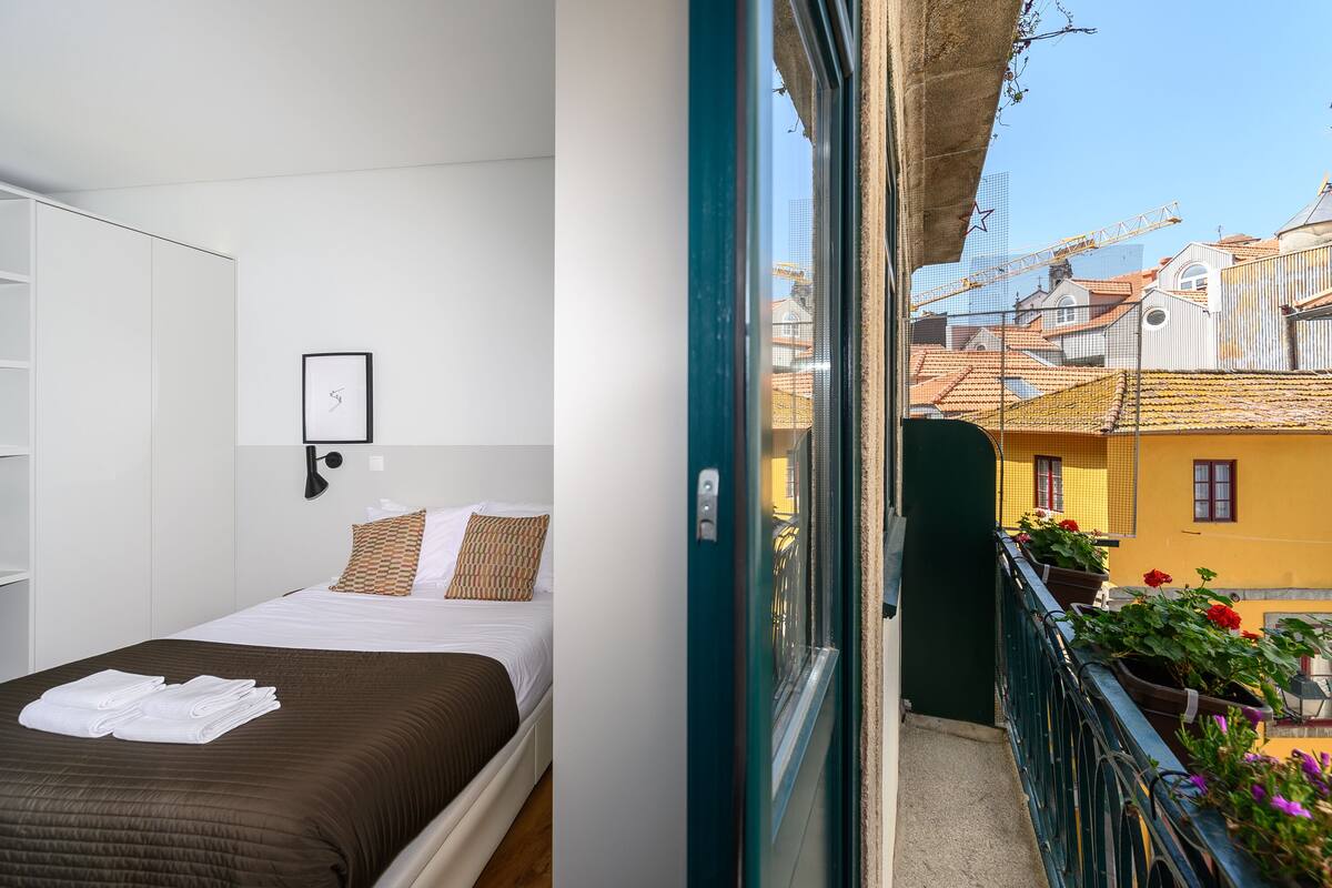 A hotel room with a bed, a framed picture on the wall, and a balcony with potted plants and a view of the cityscape.