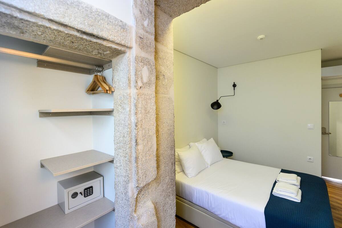 A hotel room with a bed, a bedside table, a hanging coat rack, a wall-mounted lamp, and a small safe.