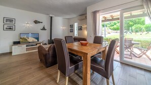 A dining area with a wooden table and chairs, a TV on a stand, and a view of the outdoors through a glass door. - C22 Quinta do paraiso Superbly Appointed and Furnished Bungalow (Carvoeiro)