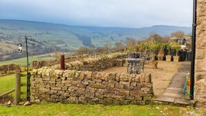 Un mur de pierres avec un portail en métal, un banc de pierre et une table avec des chaises, le tout situé dans un cadre rural.