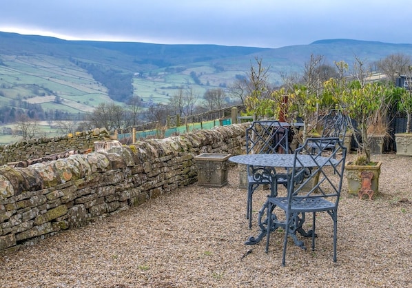 Un mur de pierre et des chaises en métal dans un jardin offrant une vue sur des collines ondoyantes.