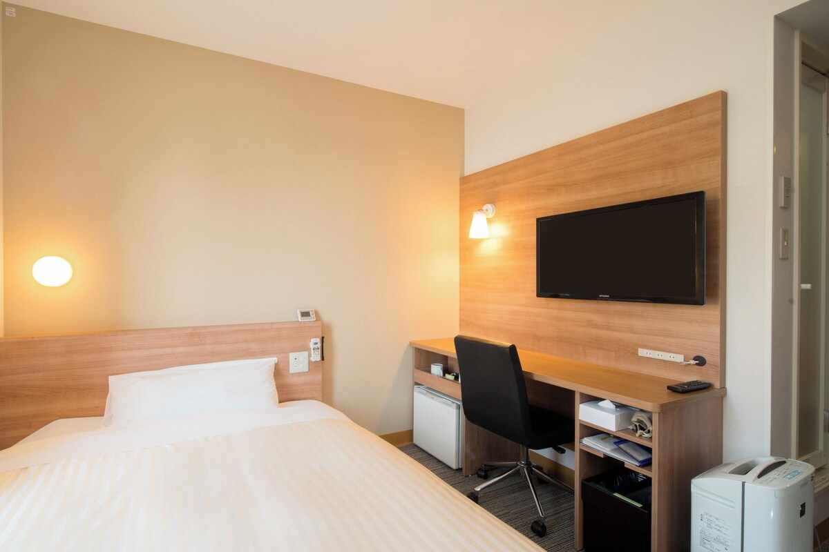 A hotel room with a bed, a desk, a chair, a television, and a small refrigerator.