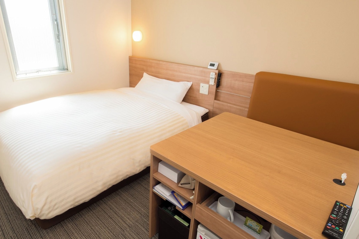 A hotel room with a single bed, a bedside table with a remote control, a heater, and a window with blinds.