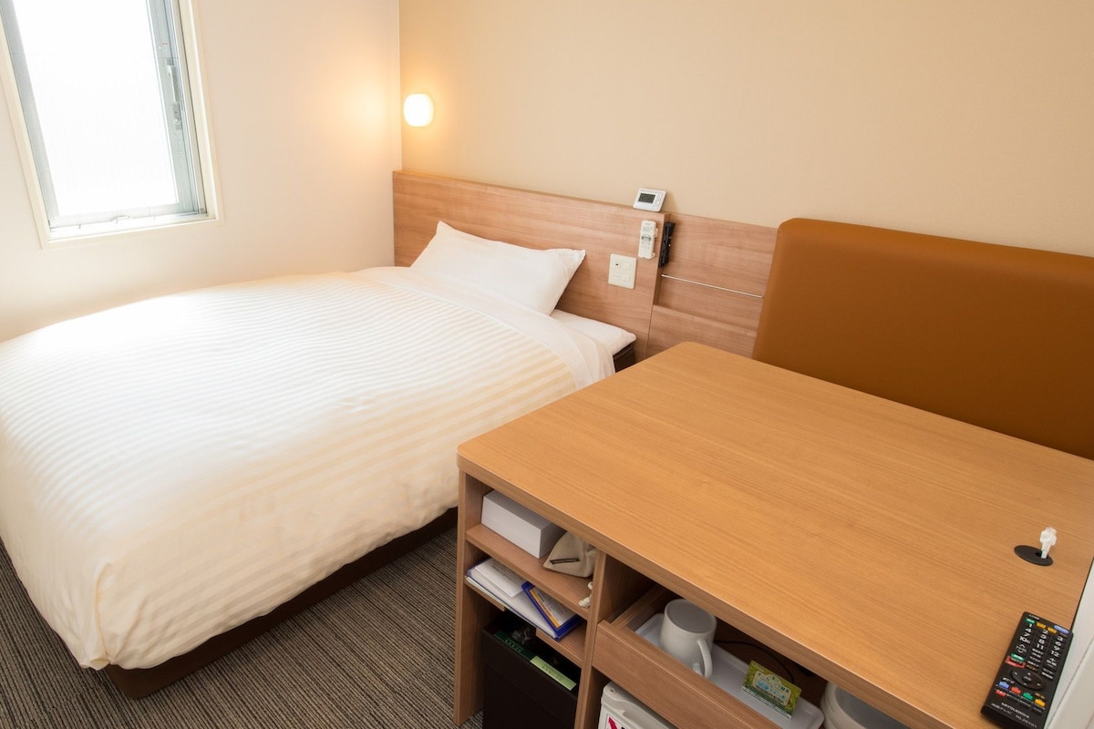 A hotel room with a bed, bedside table, and a small desk with a chair.