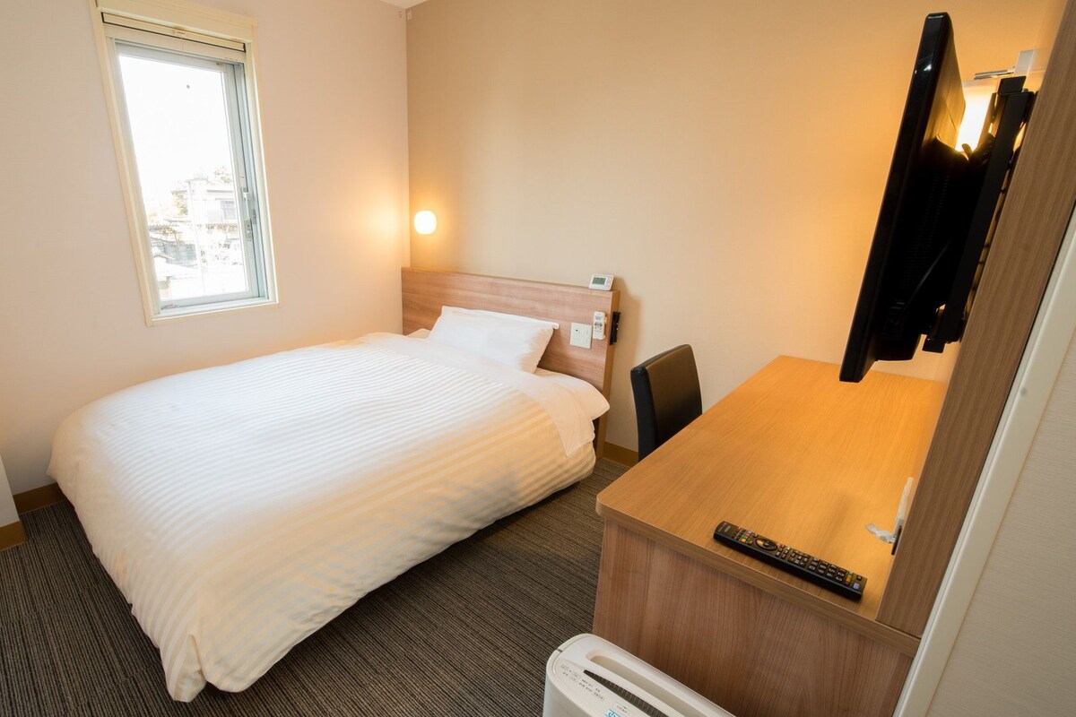 A hotel room with a bed, a desk with a chair, a television, and a window.
