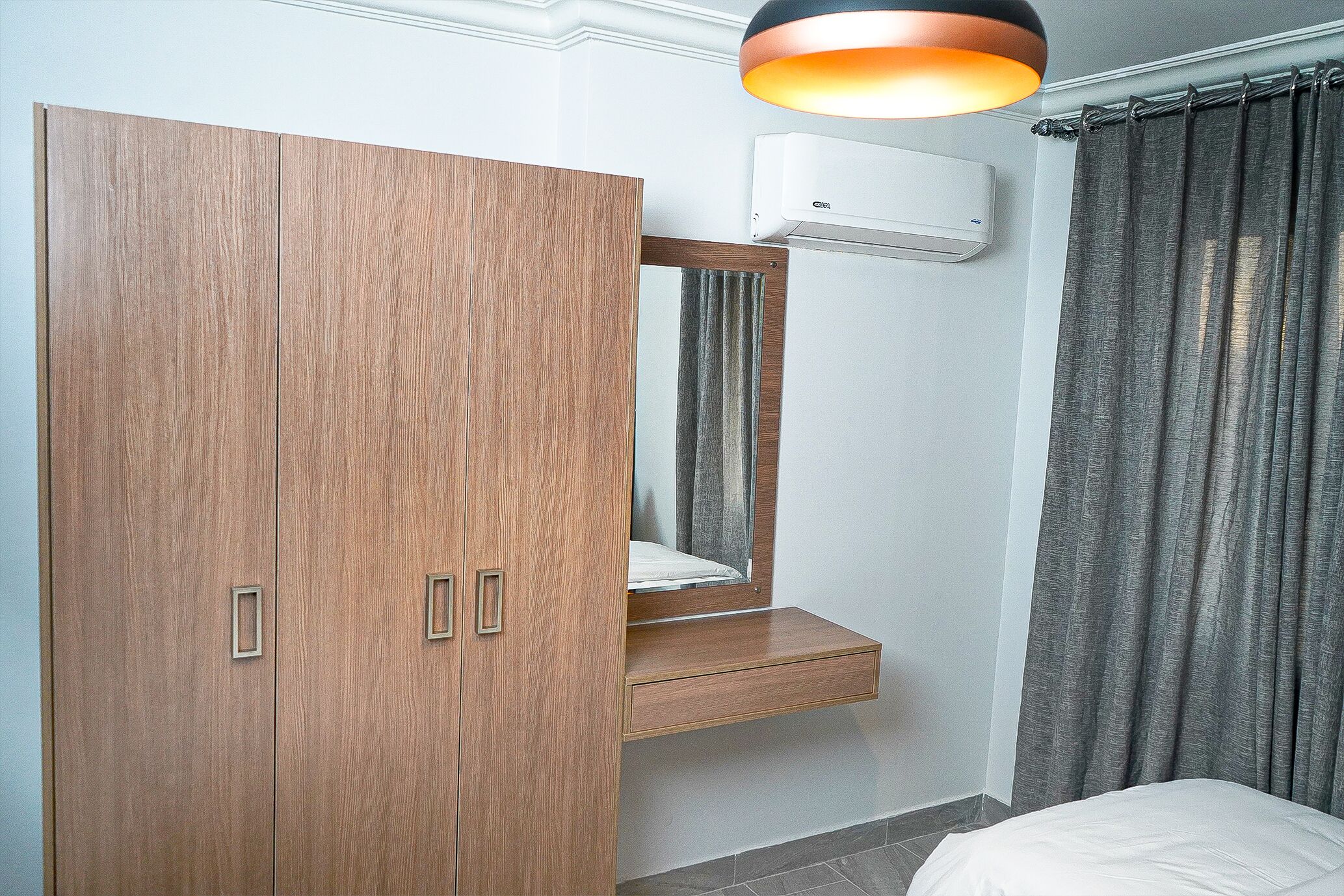 A bedroom with a wooden wardrobe, a bedside table, a mirror, and a grey curtain.